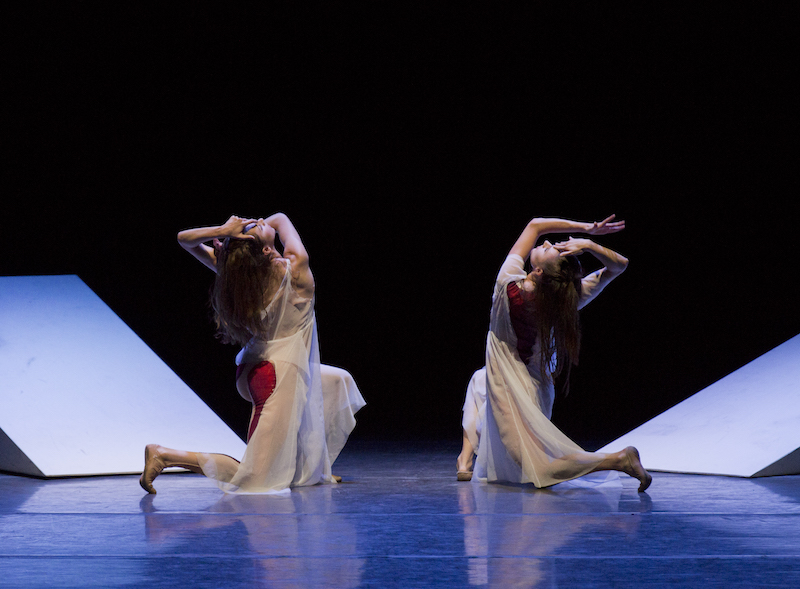 Two dancers in white sheer frocks with red on the backside kneel away from the audience. Their faces are in profile. Upstage of them are two white triangular ramps.